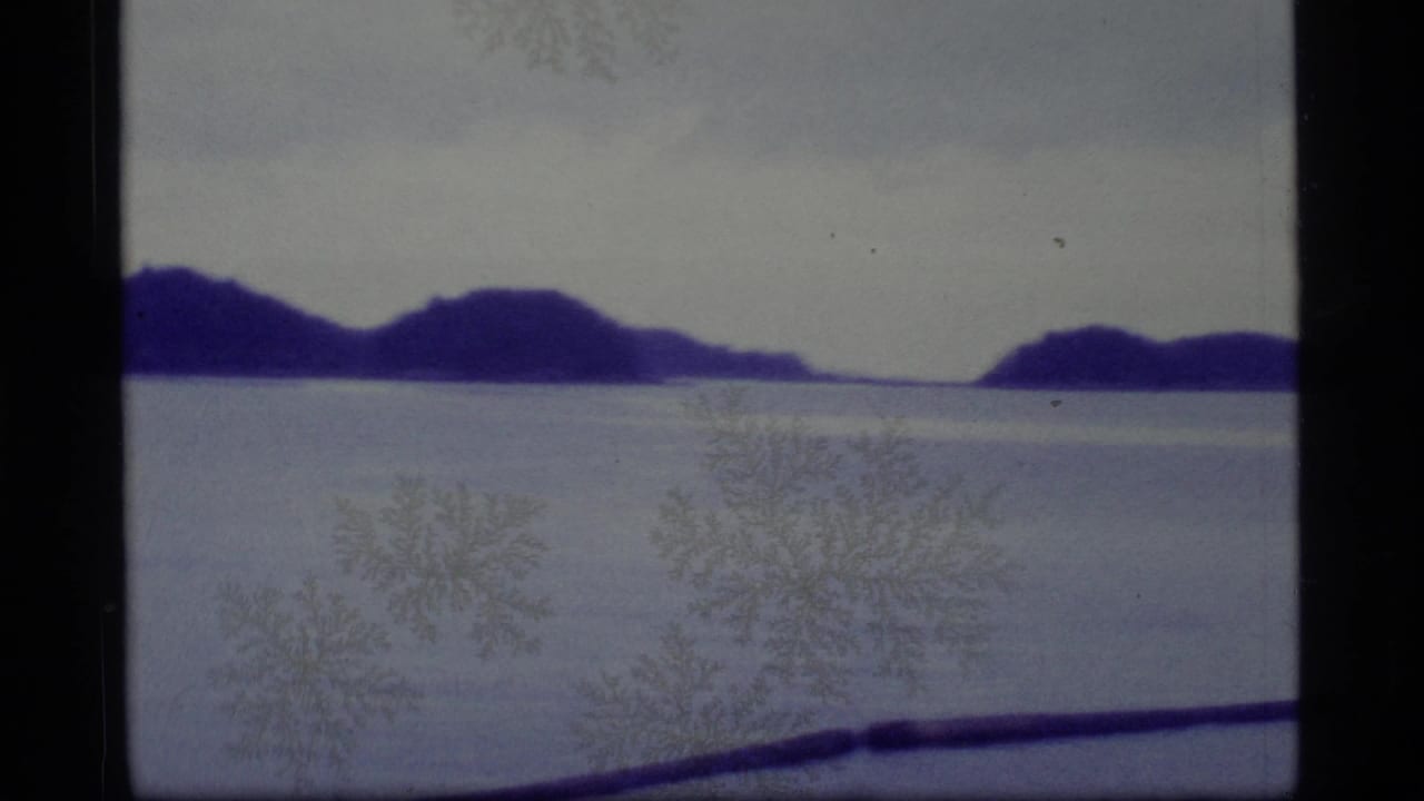 1977 Alaska 8mm frame showing distant island silhouettes beyond a pale frozen or watery expanse with film marks visible.