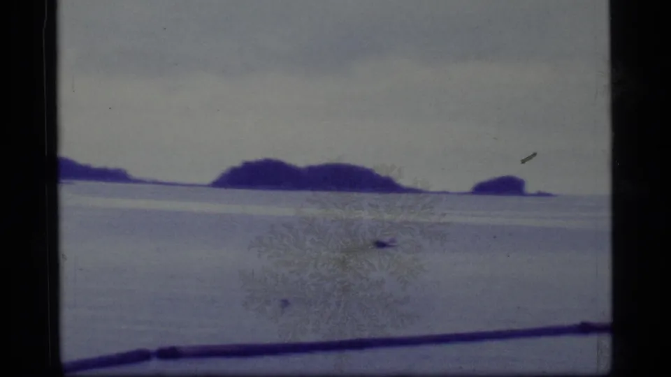 1977 Alaska 8mm view of low islands across a pale expanse, with blue-purple color shift and surface film marks.