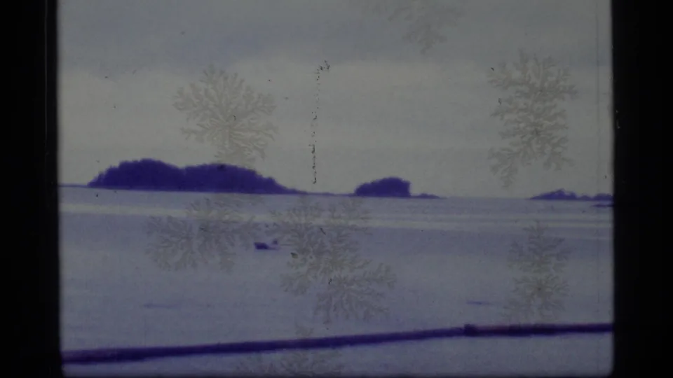 1977 Alaska home-movie frame showing a small craft near the horizon, distant islands, and a dark boat rail in the foreground.
