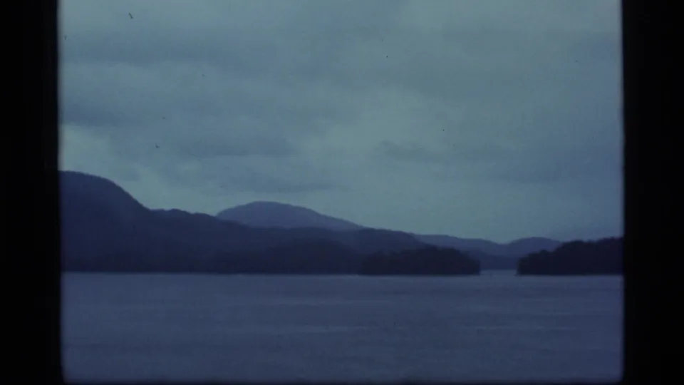 1977 Alaska 8mm frame showing calm sea, low wooded shoreline, and distant mountains under a gray-blue overcast sky.