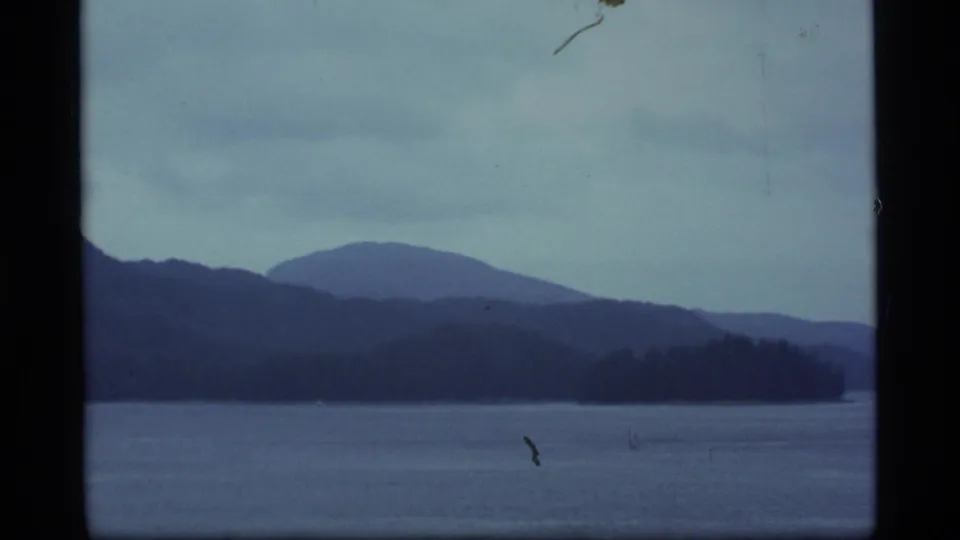 1977 Alaska 8mm frame showing open water, a dark wooded shoreline, rounded distant mountains, and light surface film marks.