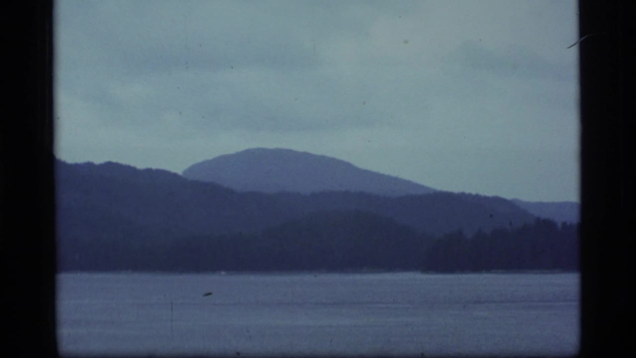 1977 Alaska 8mm frame showing dark water, distant wooded shoreline, and layered blue mountain silhouettes beneath a grey sky.