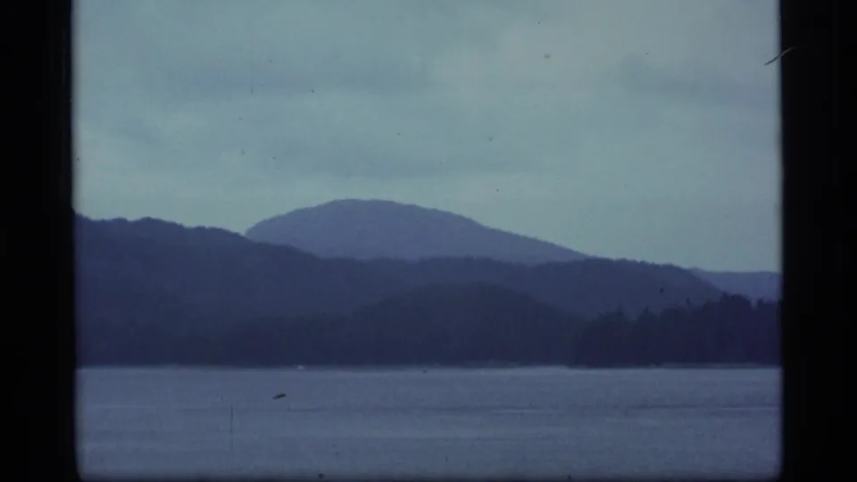 1977 Alaska 8mm frame showing dark water, distant wooded shoreline, and layered blue mountain silhouettes beneath a grey sky.