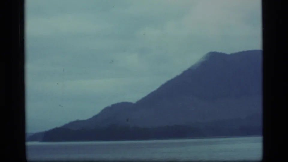 1977 Alaska 8mm frame with a broad mountain silhouette rising beyond calm water and a low, cloudy horizon.