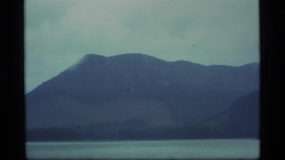 1977 Alaska 8mm frame showing soft blue mountain ridges, a narrow waterline, and visible dust in the film image.