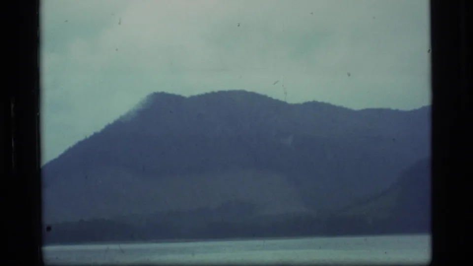 1977 Alaska 8mm frame with a broad mountain ridge, muted water, cloudy sky, and visible scratches across the scan.