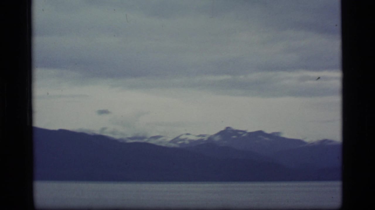 1977 8mm Alaska footage showing distant snow-streaked mountains beyond calm bay water under layered clouds.