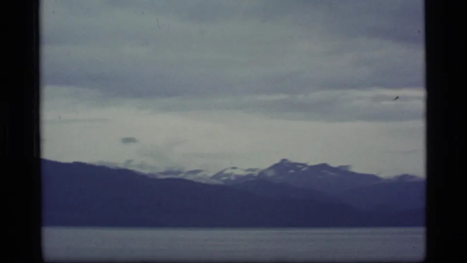 1977 8mm Alaska footage showing distant snow-streaked mountains beyond calm bay water under layered clouds.