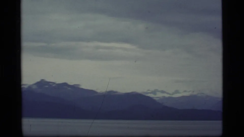 1977 8mm Alaska view of snow-capped peaks layered behind dark ridgelines across a gray-blue bay.