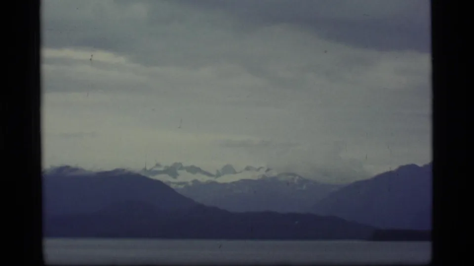 1977 Alaska home-movie frame with dark low hills, distant snowy mountains, calm water, and a heavy overcast sky.