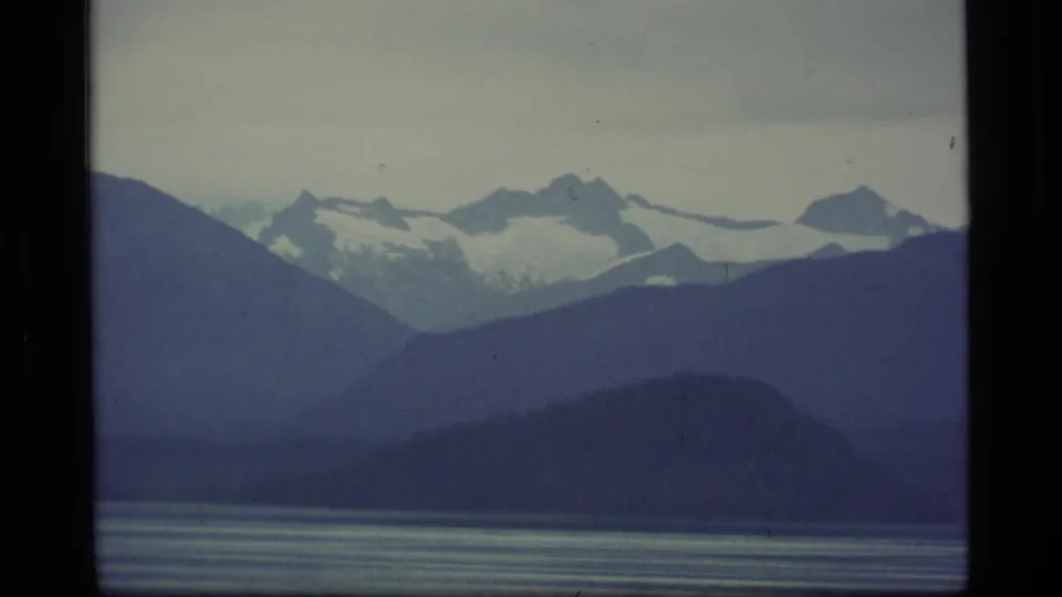 1977 8mm Alaska frame showing rugged snowy peaks beyond dark mountain slopes and lightly rippled bay water.