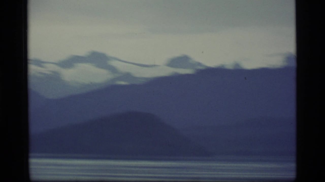 1977 8mm Alaska landscape view with layered dark ridges, snow-topped mountains, and still bay water beneath a pale sky.
