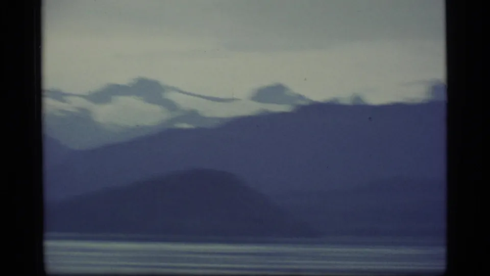 1977 8mm Alaska landscape view with layered dark ridges, snow-topped mountains, and still bay water beneath a pale sky.