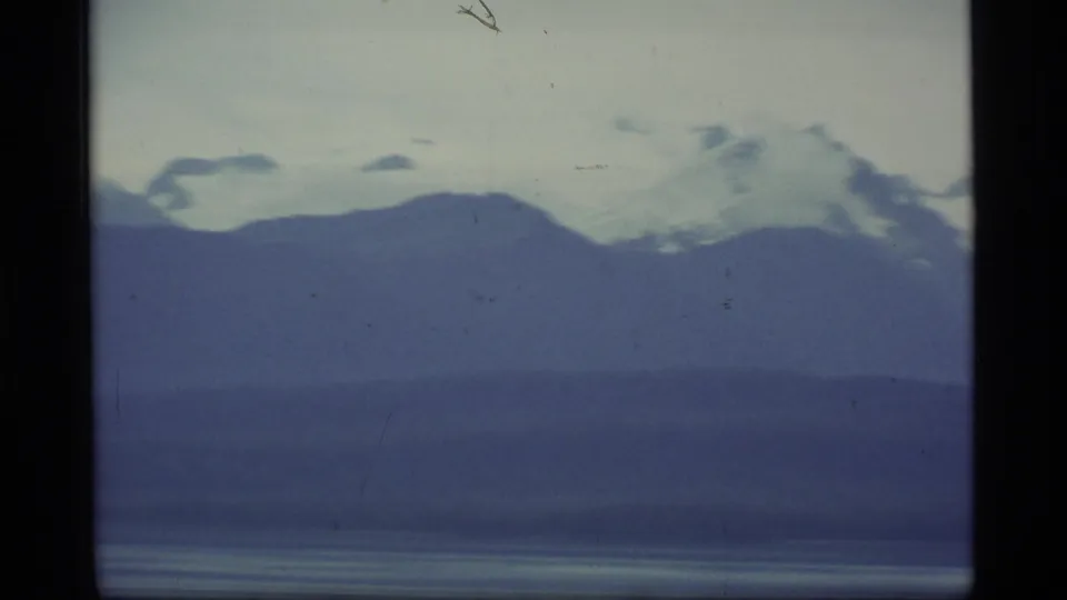 A distant Alaska bay landscape shows dark mountain layers, snow patches on high peaks, and muted blue 8mm exposure.