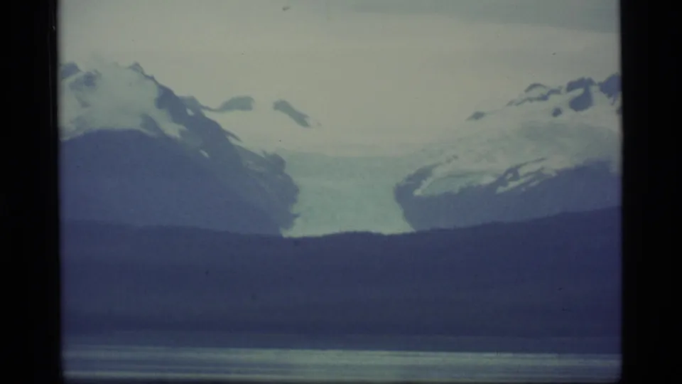 1977 Alaska home-movie frame showing a pale glacier descending between dark slopes, with water and snowy mountains nearby.