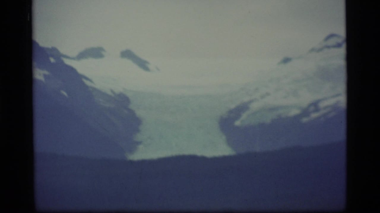 1977 Alaska 8mm frame showing distant snow-covered mountains above a dark ridge and muted sky.