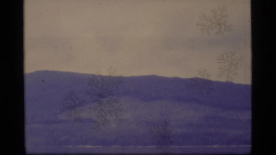1977 Alaska home-movie frame of layered hills across water or low land, marked by 8mm grain and snowflake-like film artifacts.