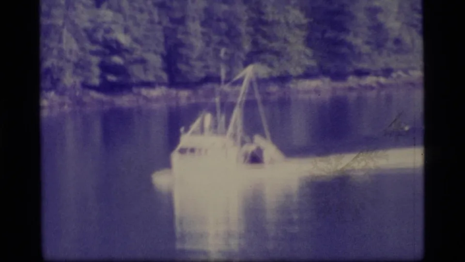 1977 Alaska 8mm frame showing a small cabin boat moving across a calm lake before a dense evergreen shoreline.