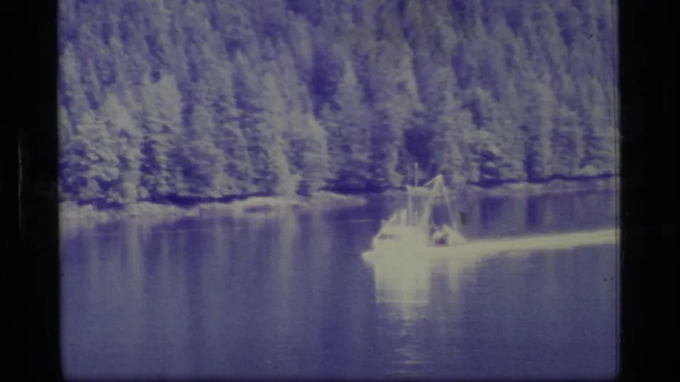 1977 Alaska home-movie frame with a small boat crossing calm water beneath a dense evergreen shoreline.