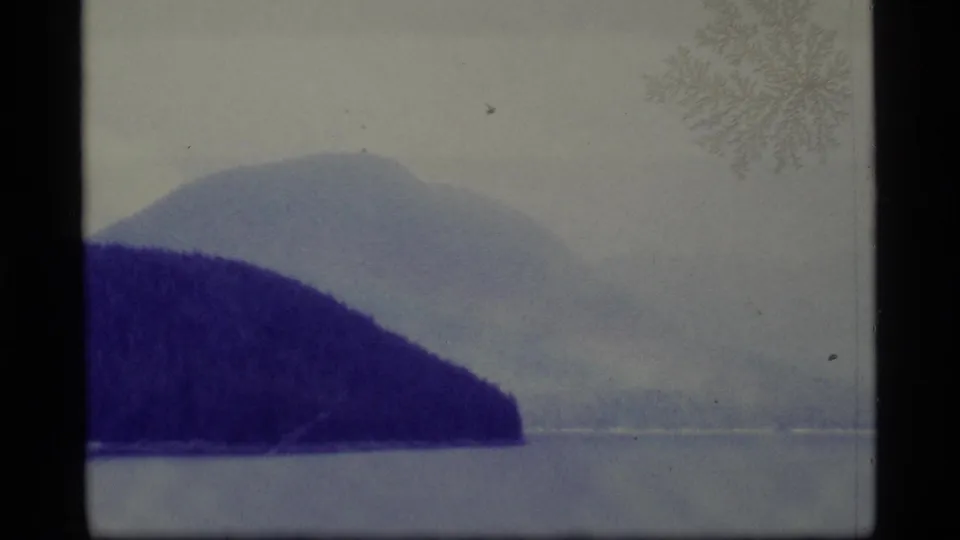 Alaska lake view on 8mm film, showing a dark forested slope, pale water, and a distant mountain form under hazy light.