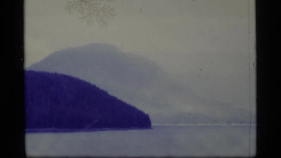 1977 Alaska 8mm frame showing a broad lake, dark shoreline, distant mountains, pale haze, and black film borders.