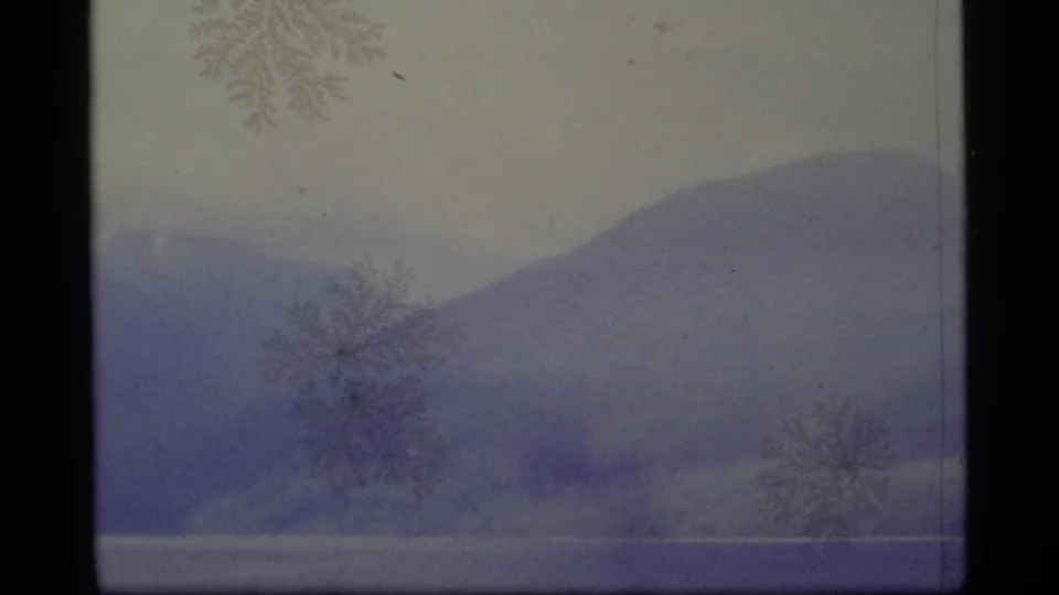 1977 Alaska 8mm frame of rounded mountains and lake water, marked by haze, blue-purple fading, and surface patterns.