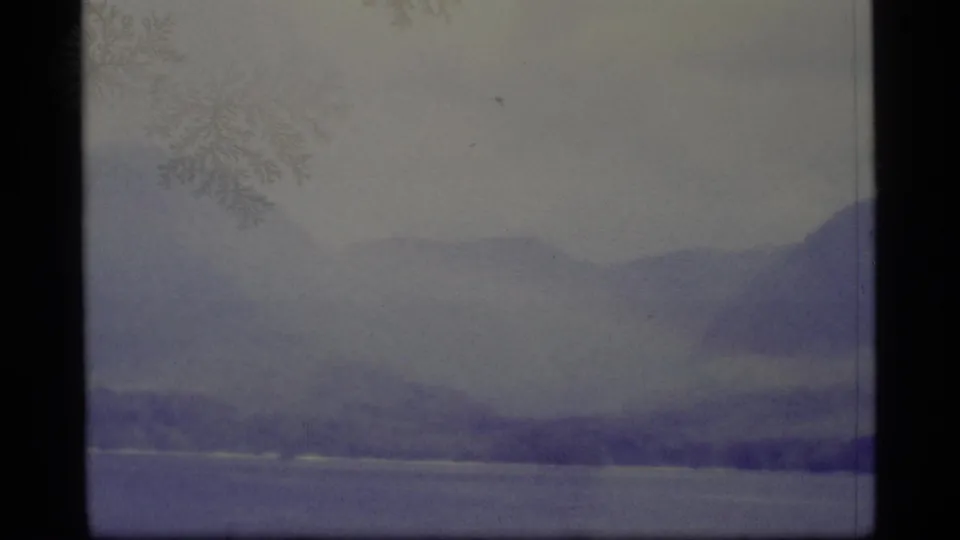 1977 Alaska 8mm frame showing low blue hills, a muted lake edge, and a dark band of shoreline trees.