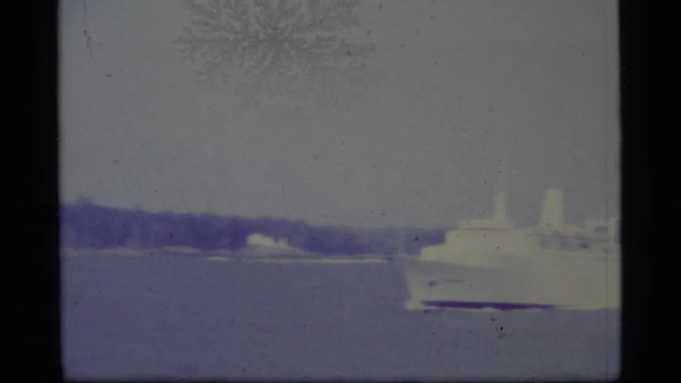 1977 Alaska 8mm still with the white cruise ship partly cropped at right, calm water in front, and a dark shoreline under muted sky.