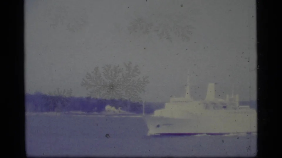 1977 Alaska home-movie frame showing a white cruise ship on calm water, distant shore, pale sky, and prominent 8mm grain and surface marks.