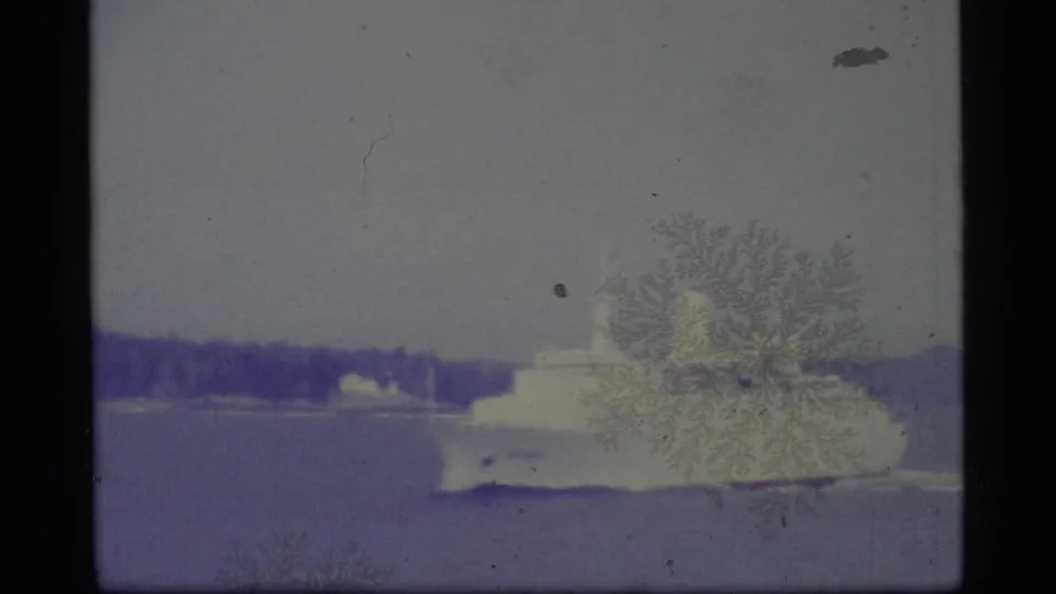 1977 Alaska 8mm frame showing a white passenger vessel on calm water, with distant shore, dark film edge, grain, and emulsion marks.