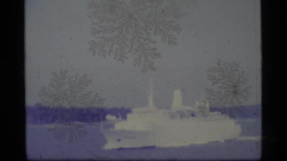 1977 Alaska home-movie frame with a white passenger vessel passing across blue water beneath a low, distant shoreline.