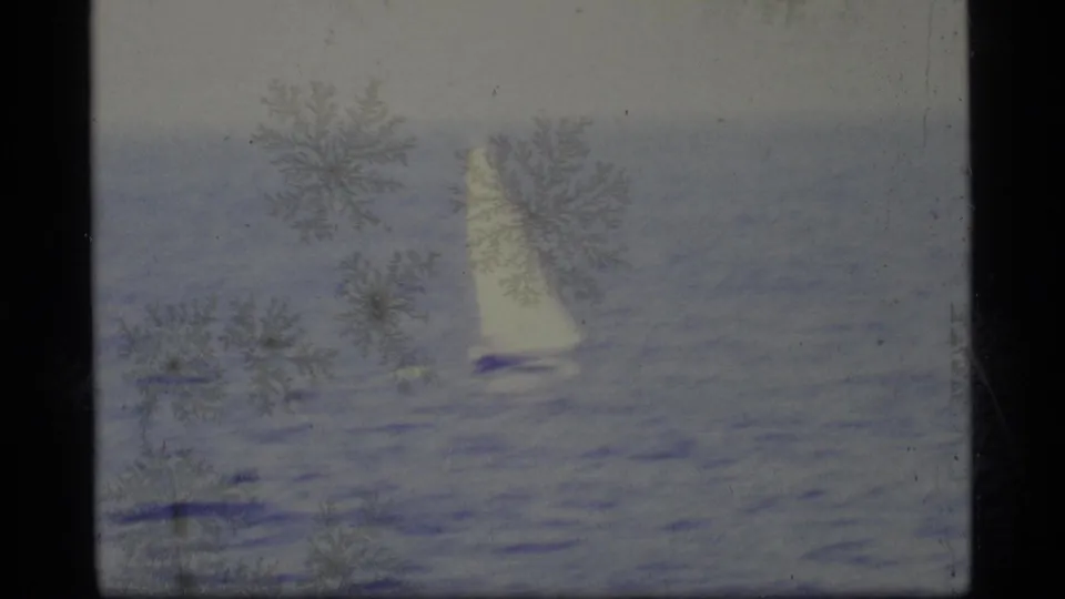 A lone white sailboat moves across calm water in 1977 Alaska home-movie footage, with visible 8mm grain and emulsion marks.