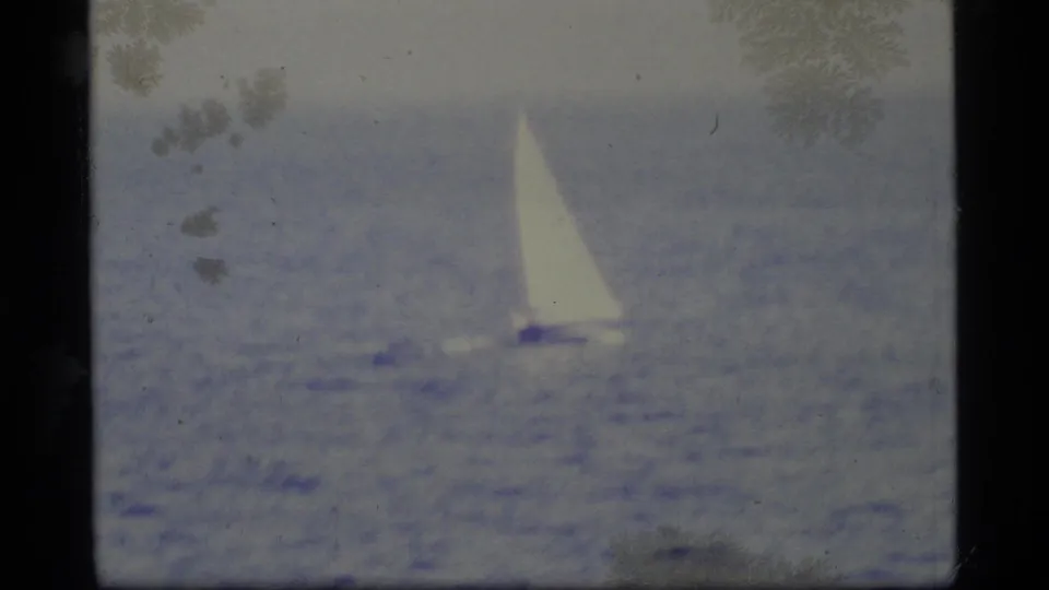 A white-sailed boat crosses calm blue water off the Alaskan coast, rendered with soft 8mm focus and visible frame wear.