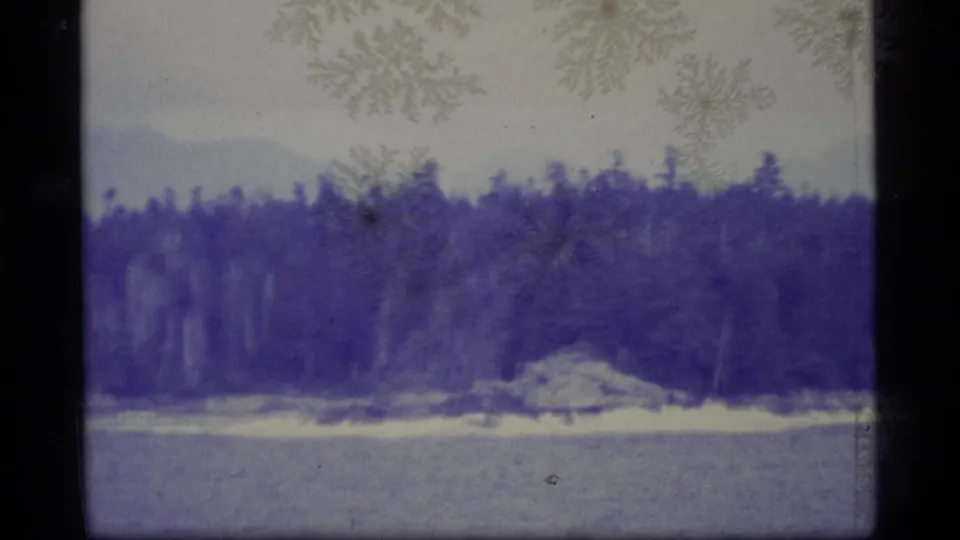1977 Alaska 8mm frame with a distant wooded shoreline, a small rocky outcrop, faint mountains, and visible film wear.
