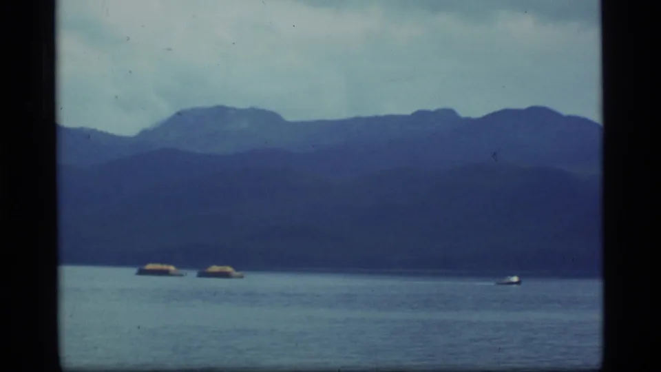 1977 Alaska 8mm frame showing open water, mountain silhouettes, distant boats, and a small airborne object above the waterline.