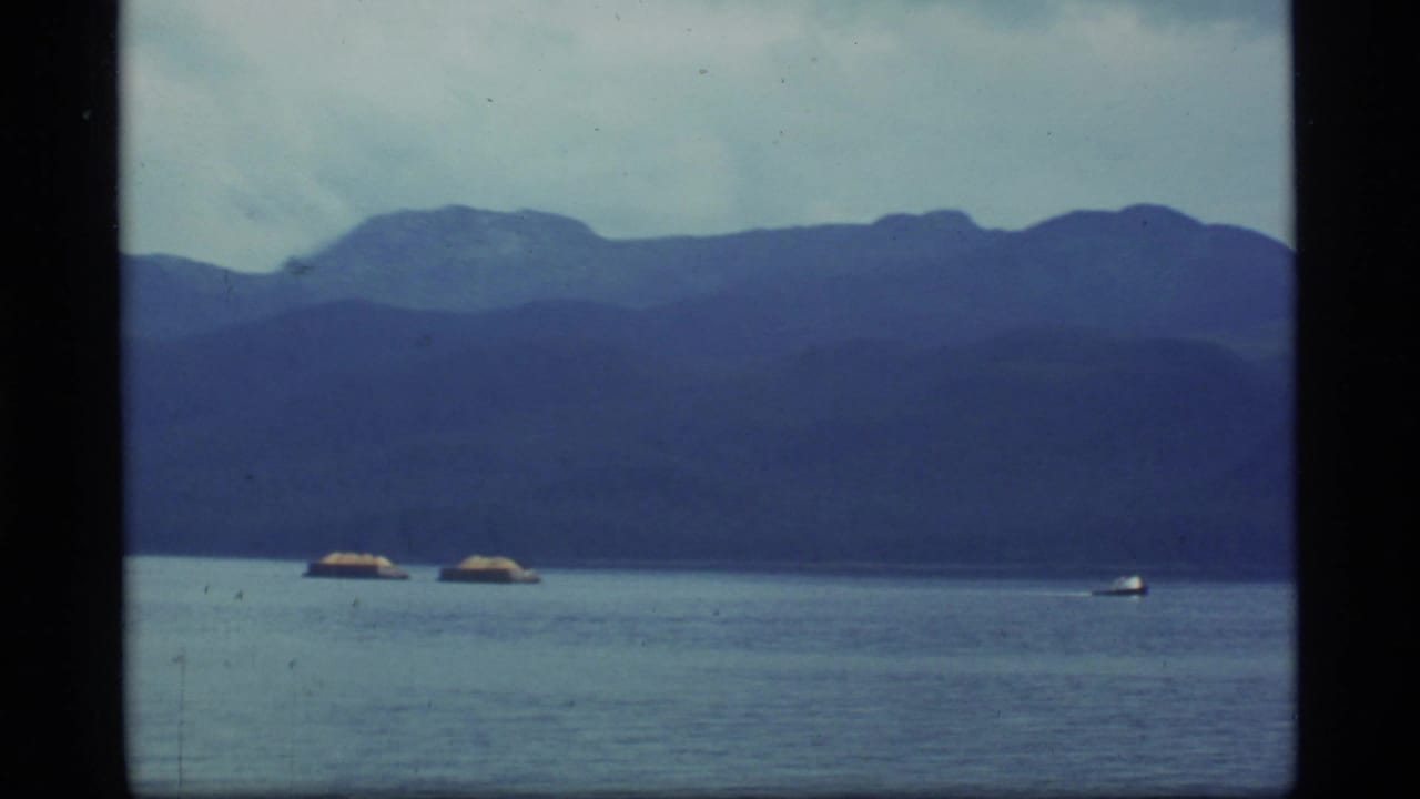1977 Alaska 8mm frame showing a calm lake, distant mountains, cloudy sky, and small boats near the horizon.