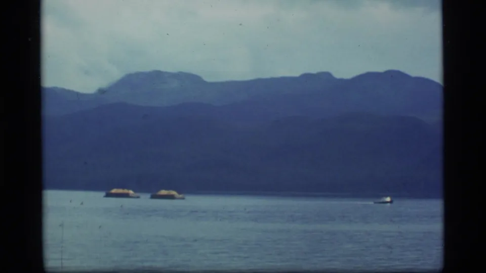 1977 Alaska 8mm frame showing a calm lake, distant mountains, cloudy sky, and small boats near the horizon.