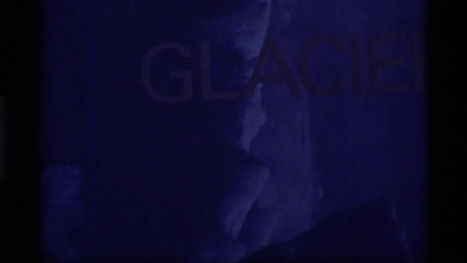 1977 Glacier Bay 8mm frame showing a dim blue title-card view with faint GLACIER lettering, grain, and dark film edges.