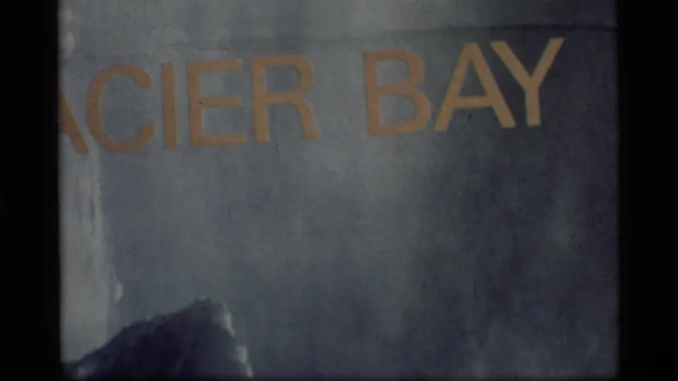 Close projected Glacier Bay title lettering from 1977 Alaska travel footage, with grain, muted color, and frame-edge darkness.