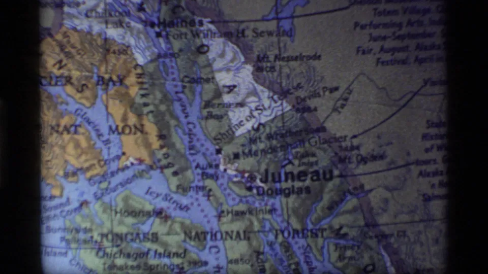 1977 Alaska travel frame showing a printed map with Glacier Bay and Juneau labels, waterways, terrain marks, grain, and color shift.