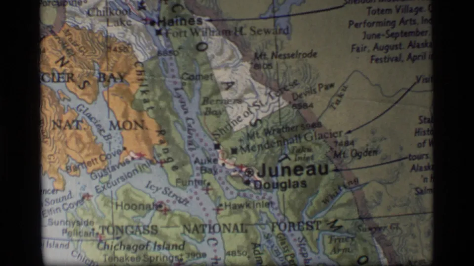 Grainy 1977 home-movie frame of a printed Alaska map showing Juneau, Douglas, and Tongass National Forest text.