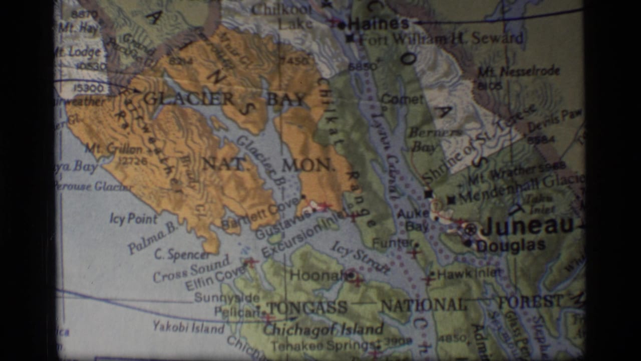 1977 8mm frame of a printed Alaska map centered on Glacier Bay and Juneau with coastal inlets and mountain shading.