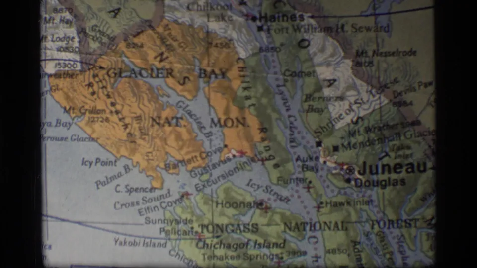 1977 8mm frame of a printed Alaska map centered on Glacier Bay and Juneau with coastal inlets and mountain shading.
