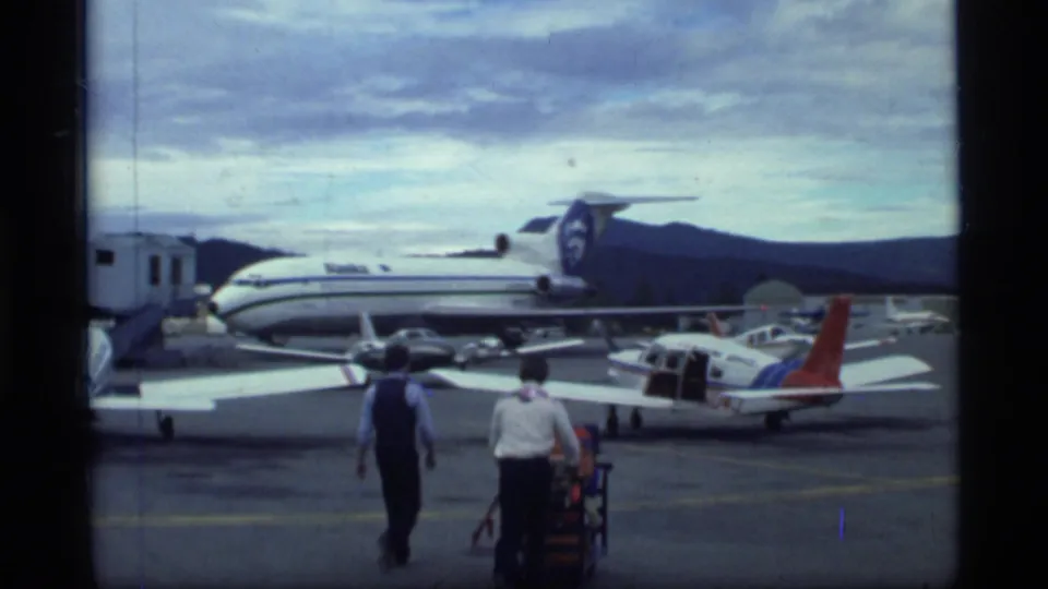 1977 Alaska travel footage shows an airport tarmac with small propeller aircraft, a larger jet, two men and luggage carts under cloudy sky.