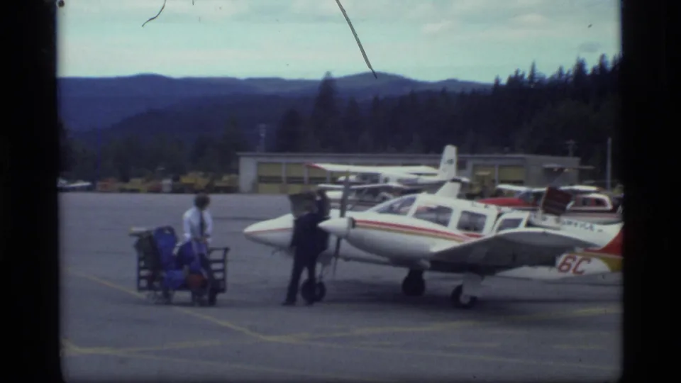 Airport tarmac scene in Alaska with a small propeller plane, two men beside luggage carts, low buildings and forested mountains.