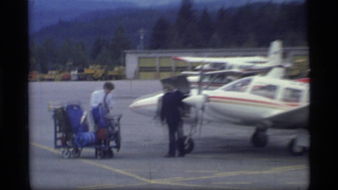 At Glacier Bay, Alaska in 1977, people and loaded luggage carts appear near a small white single-engine aircraft on the airport apron.