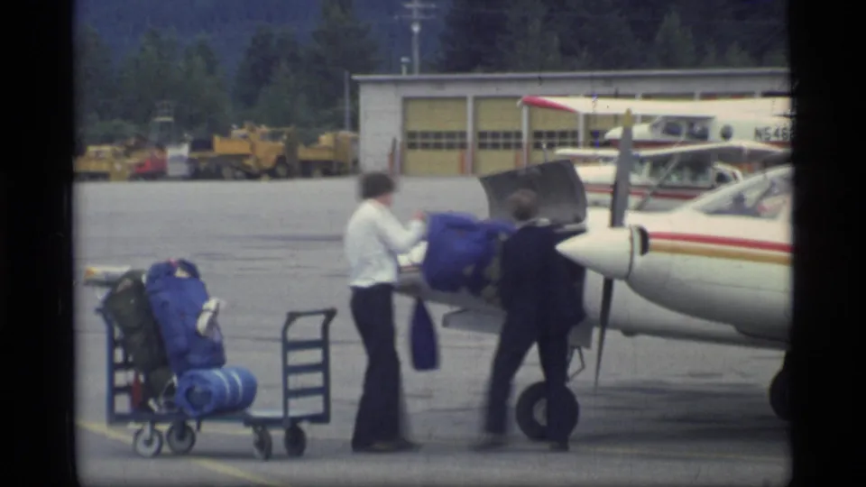 Two men handle a blue travel bag beside a small aircraft at the Glacier Bay airport, with carts and a hangar behind them.