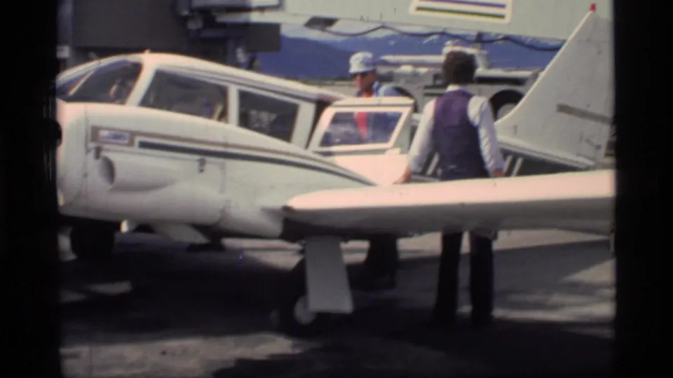 People in 1970s attire stand around a small white aircraft with an open door during flight preparation in Glacier Bay, Alaska.