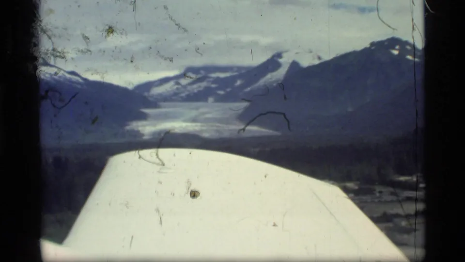 Aerial 8mm view over Glacier Bay, Alaska, with part of the aircraft visible above glacier, water, and snow-capped mountains.
