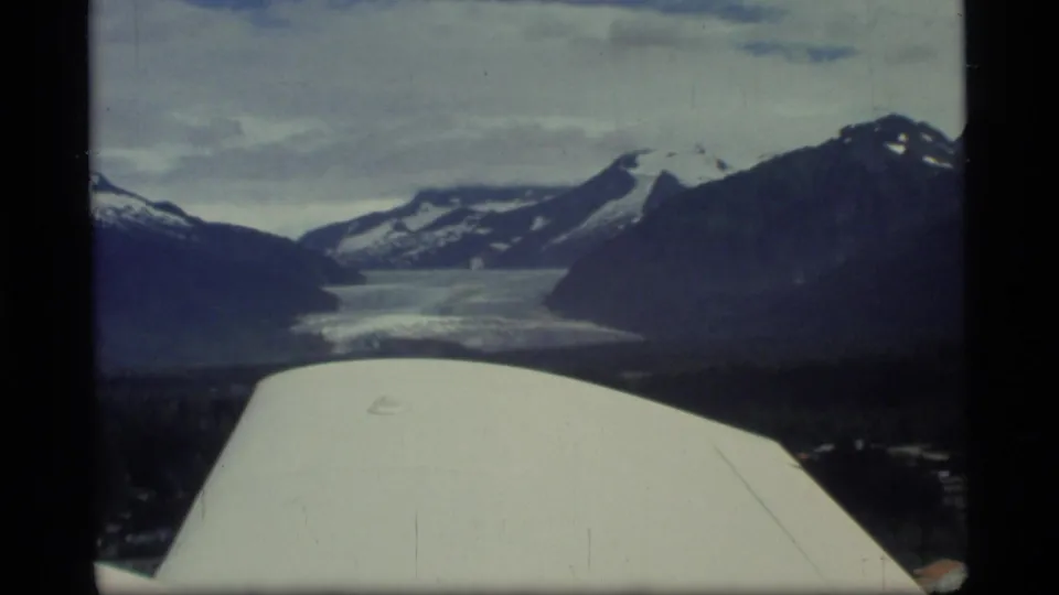 Aerial home-movie frame from Glacier Bay, Alaska, with aircraft structure in front of a glacier, valley, and mountain slopes.